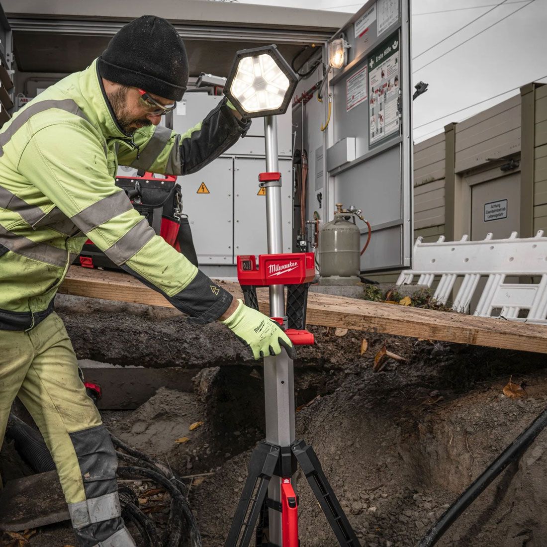 A worker adjusting the MILWAUKEE® M18 LED Stand Light on a jobsite, showcasing its adjustable head, extendable mast, and bright TRUEVIEW™ lighting in a rugged work environment.